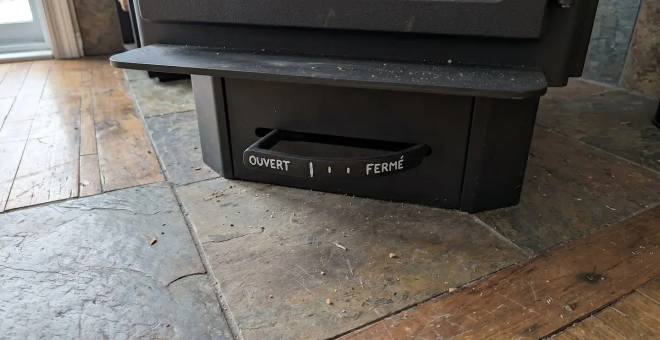 Gros plan sur la manette de réglage de l'arrivée d'air située sous un poêle à bois contemporain, avec indication des positions ouvert et fermé
